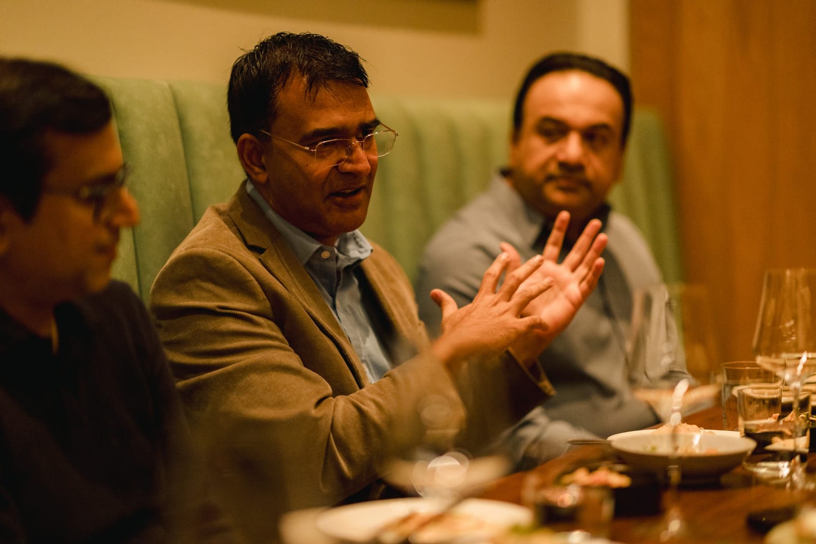 An engineering leader in a corduroy jacket gestures while making a point at the dinner table, with two other guests listening
